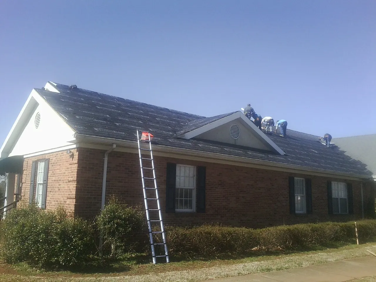 Crew tearing shingles off a brick ranch roof with a ladder set — Metal Roof Repair work by our crew in Harvard