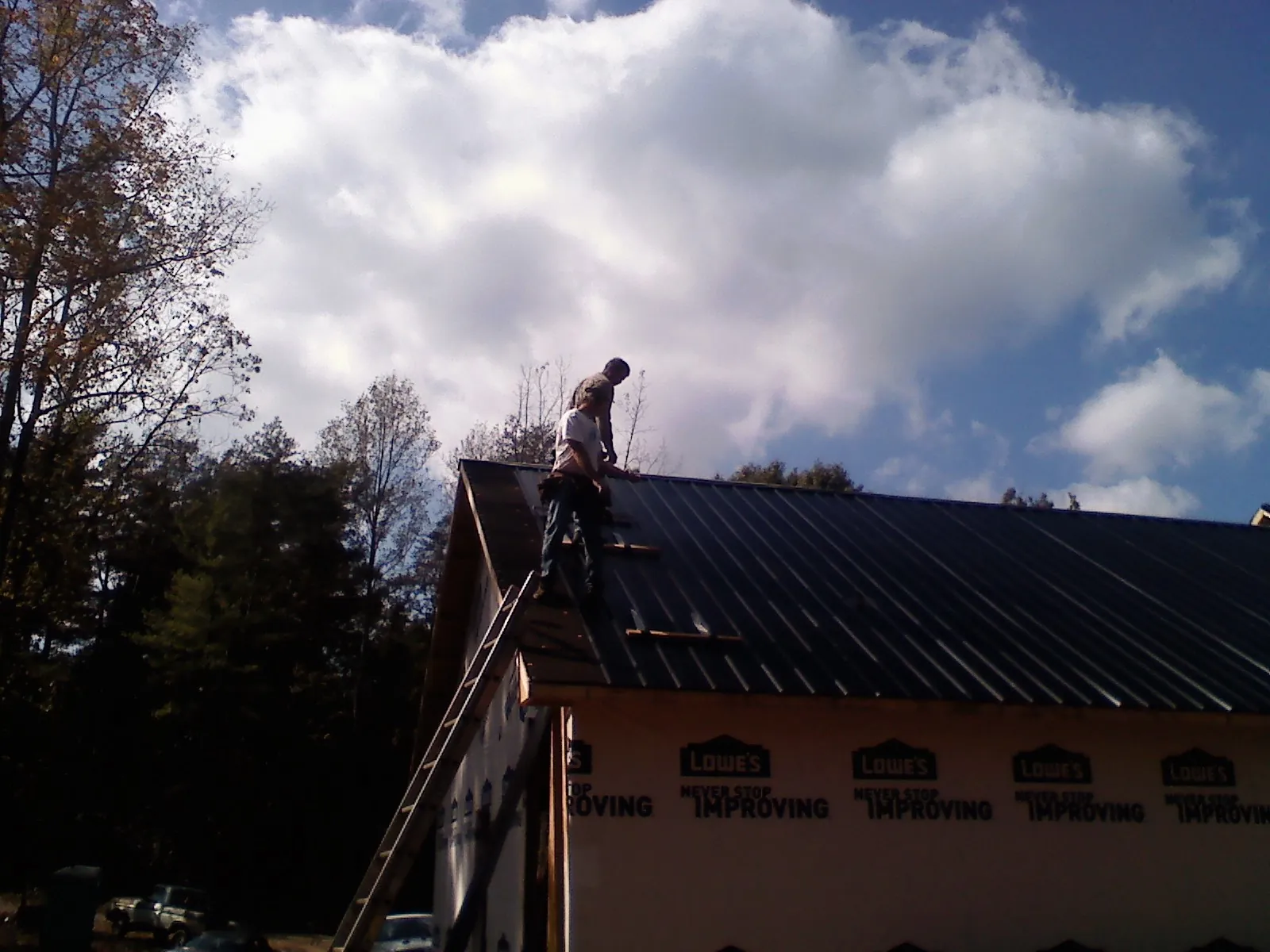 Roofer fastening dark metal panels high against a cloudy sky, from a Roof Inspection job in Harvard